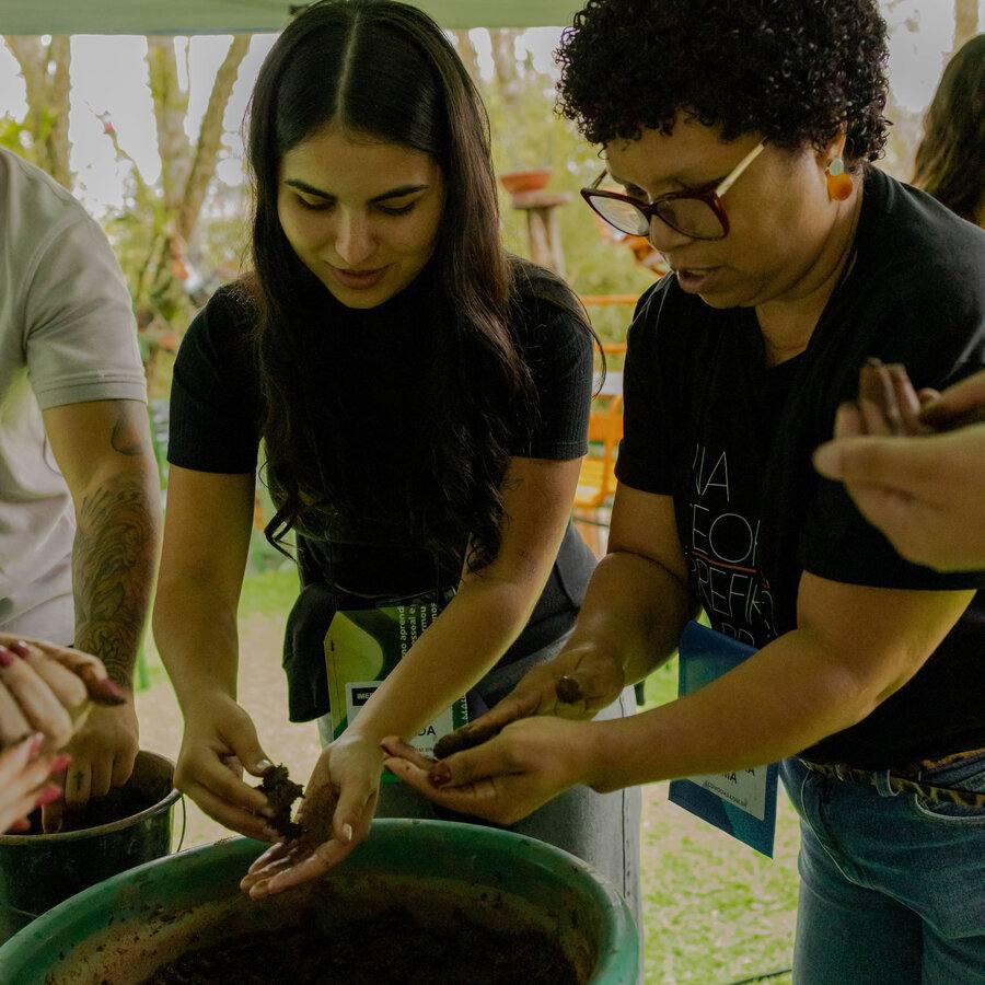 Duas participantes misturando barro e composto orgânico em uma bacia durante oficina de bombas de sementes
