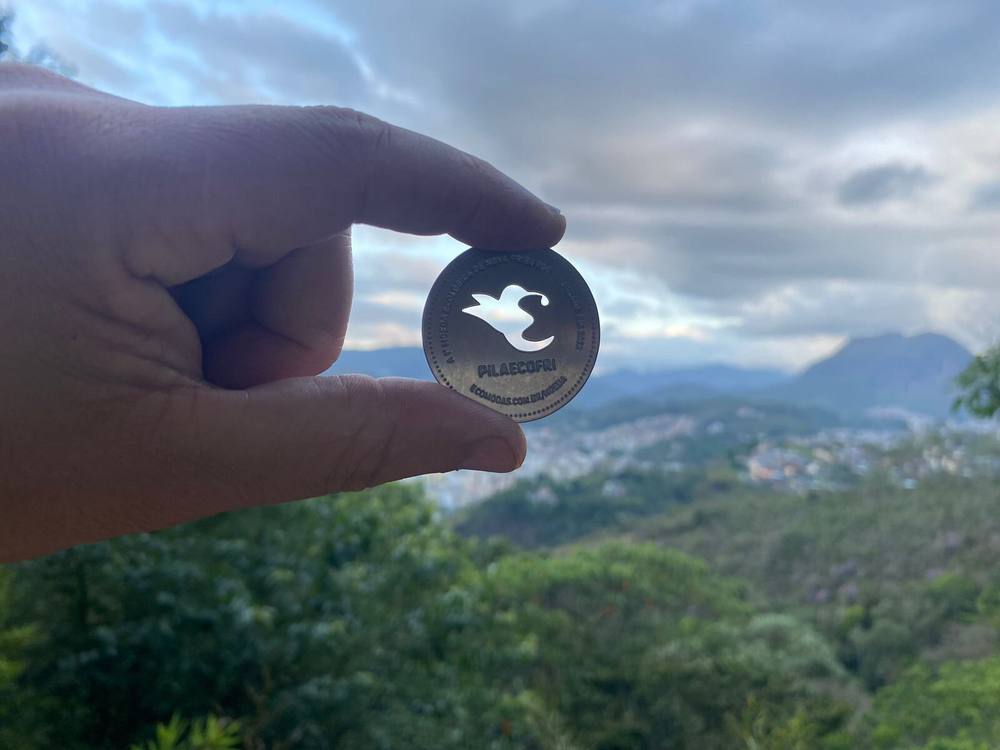 Moeda PilaEcoFri em destaque, com vista da Região Serrana do Rio de Janeiro ao fundo