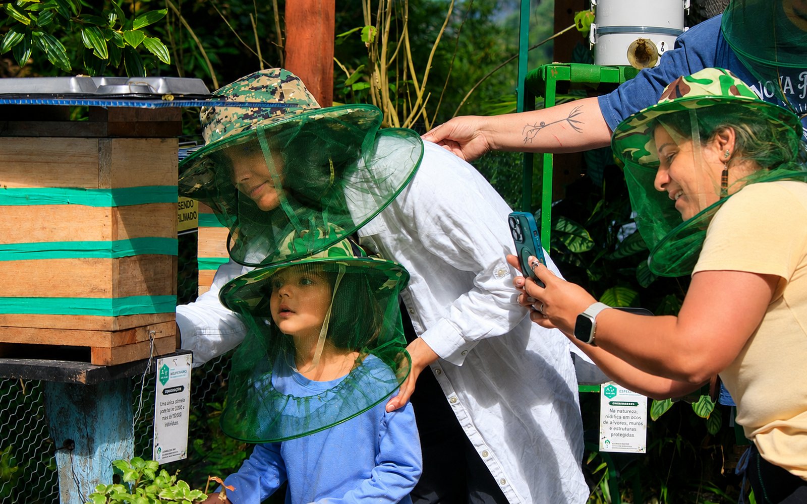 Família visitando o Meliponário Zumbido Verde, usando véus de proteção, observando uma caixa de abelhas nativas