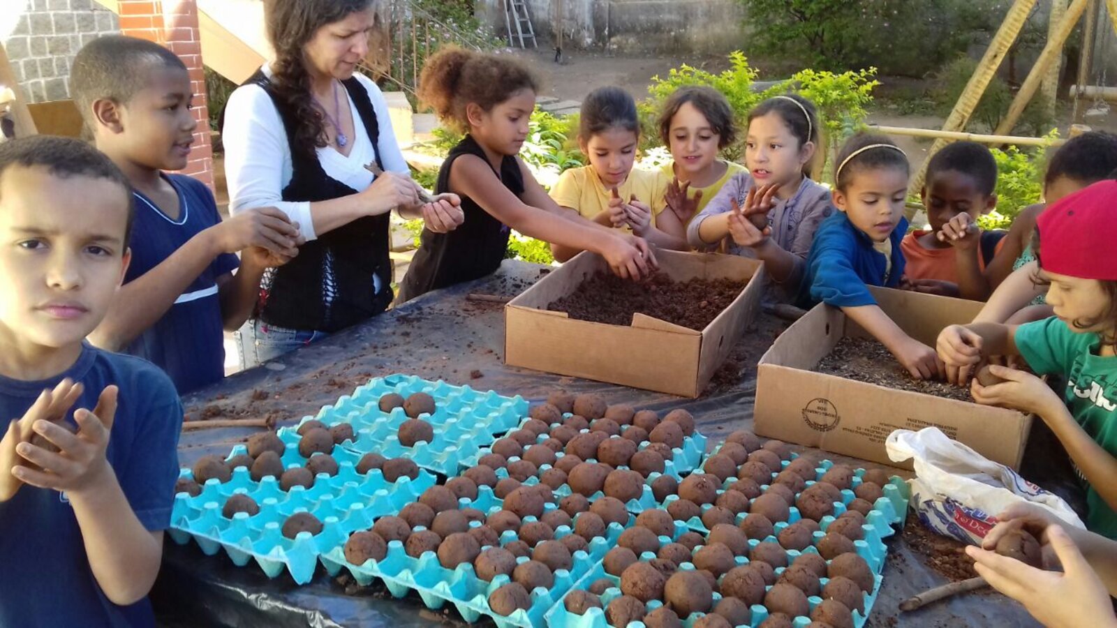 Crianças reunidas em torno de uma mesa cheia de bombas de sementes prontas, organizadas em bandejas de ovo