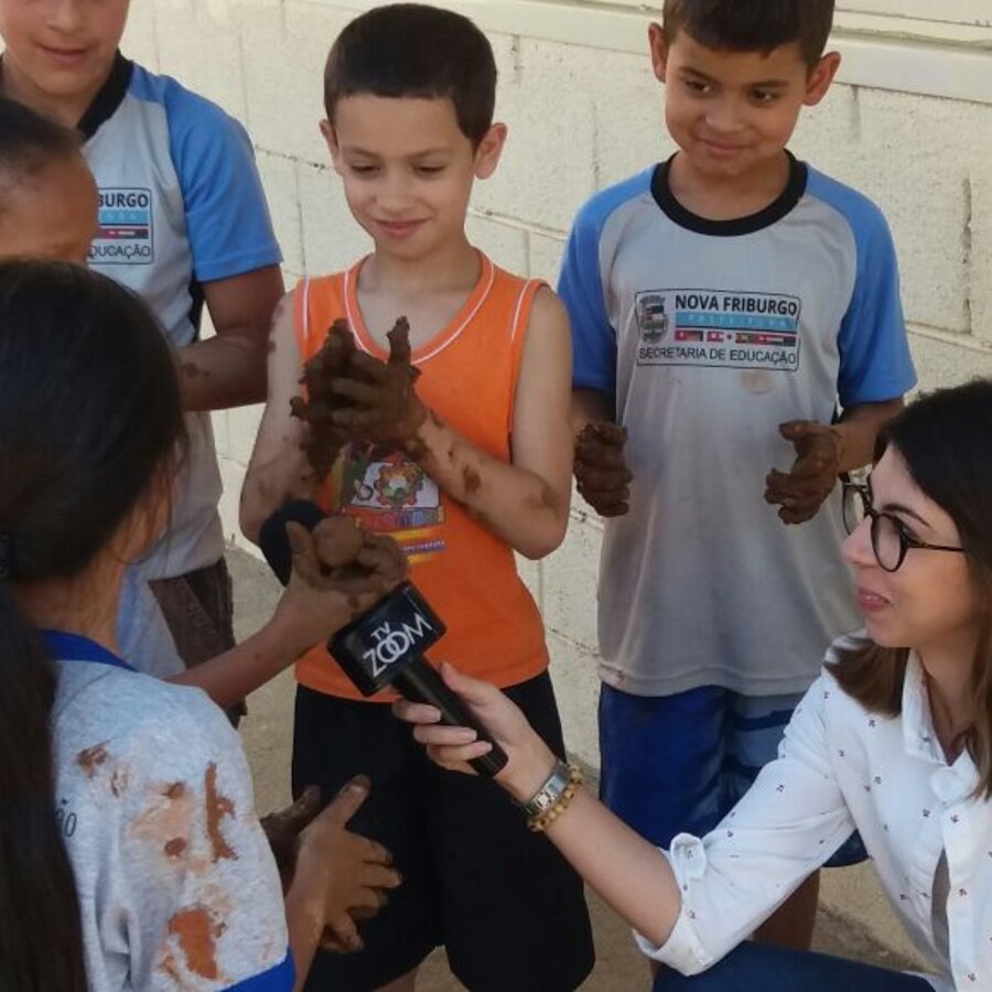 Crianças de escola municipal mostrando as mãos com barro durante entrevista para a TV local sobre o projeto