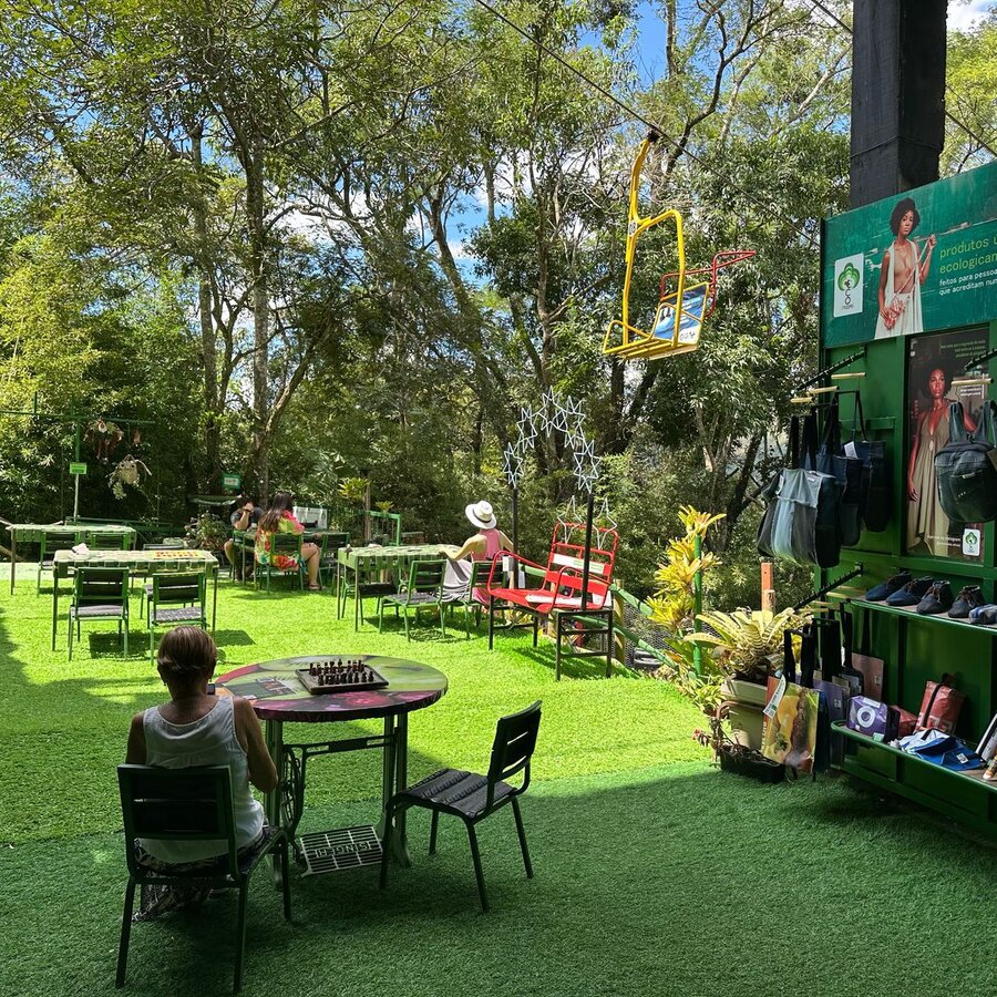 Pátio externo da EcoModas com mesas de jogos, cadeira do teleférico chegando à estação e loja ao fundo
