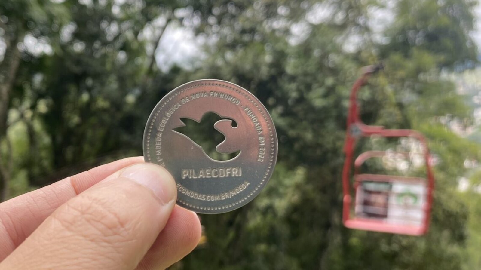 Moeda PilaEcoFri em close, com uma cadeira do teleférico de Nova Friburgo desfocada ao fundo
