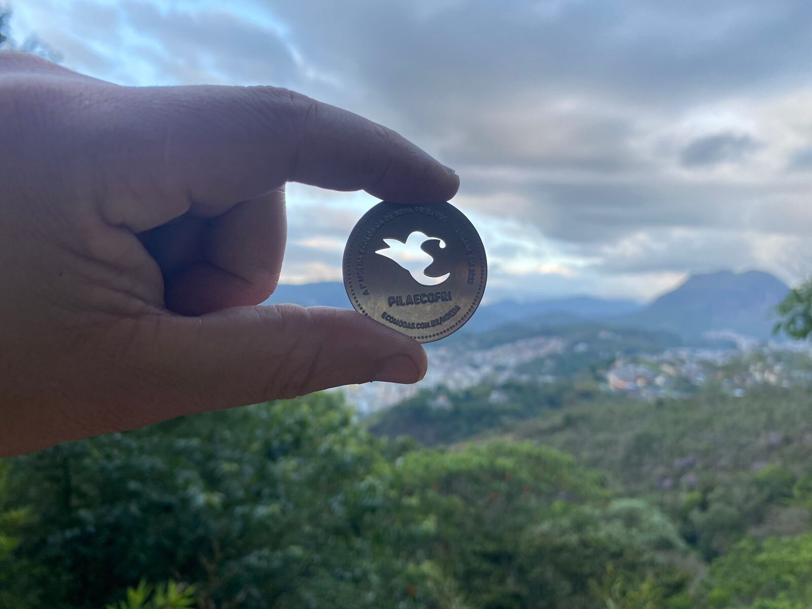 Moeda PilaEcoFri em destaque, com vista da Região Serrana do Rio de Janeiro ao fundo