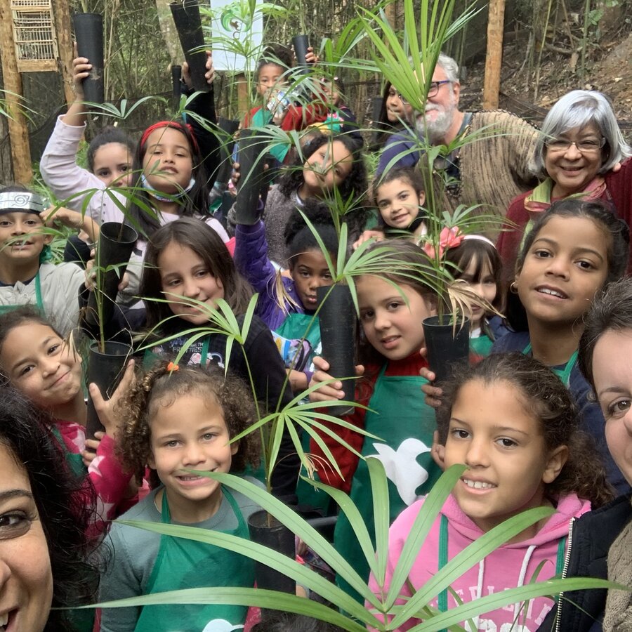 Grupo de alunos e educadores segurando mudas de palmeira em tubetes dentro do Viveiro Educandário